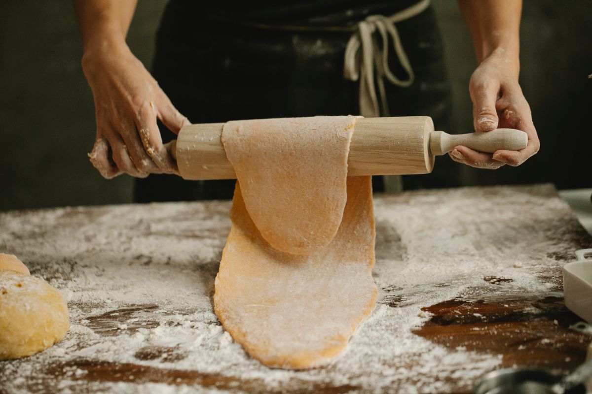 Hands rolling fresh pasta dough with a wooden rolling pin on a floured bench