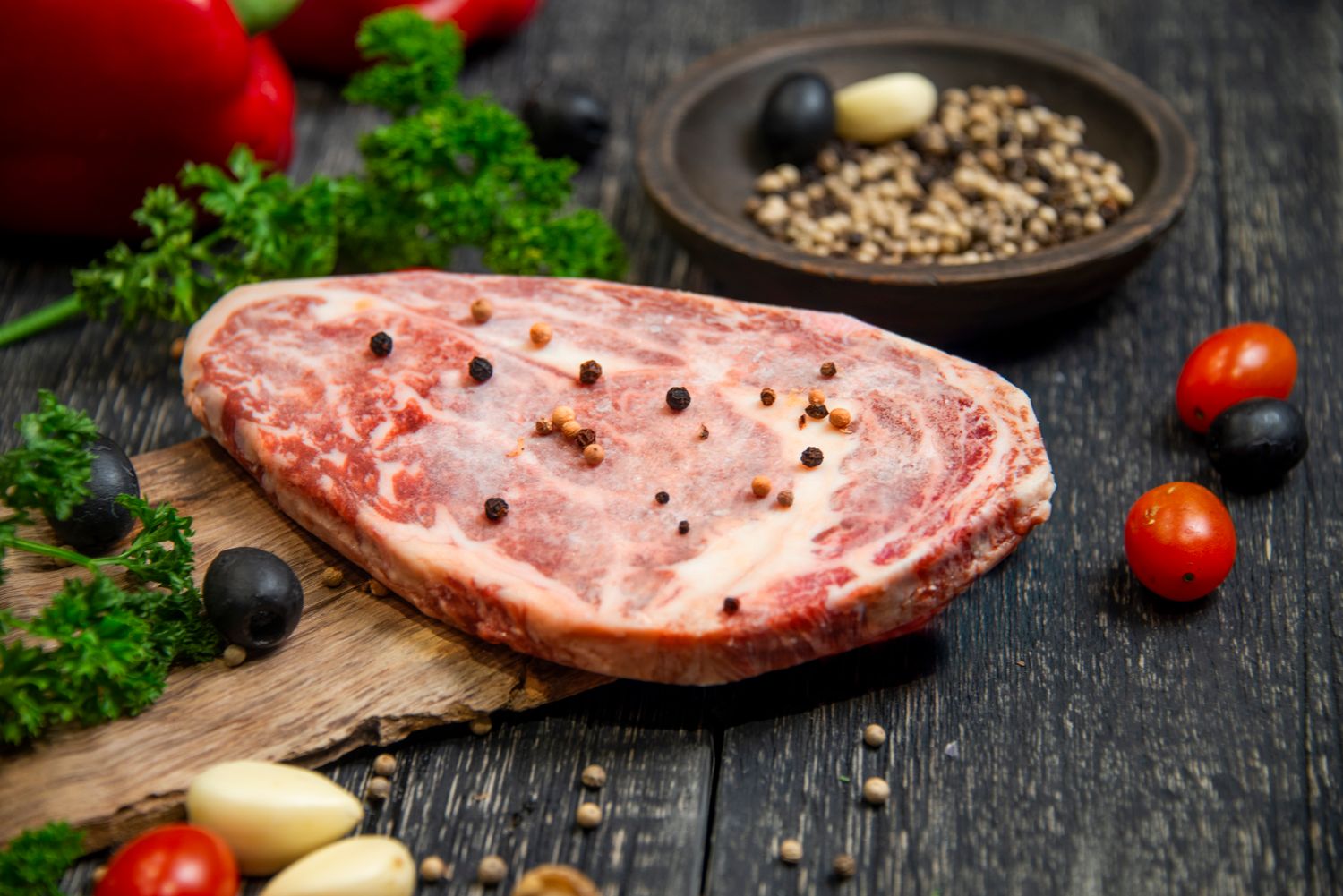 A dry-aged steak resting on a dark board with peppercorns and herbs