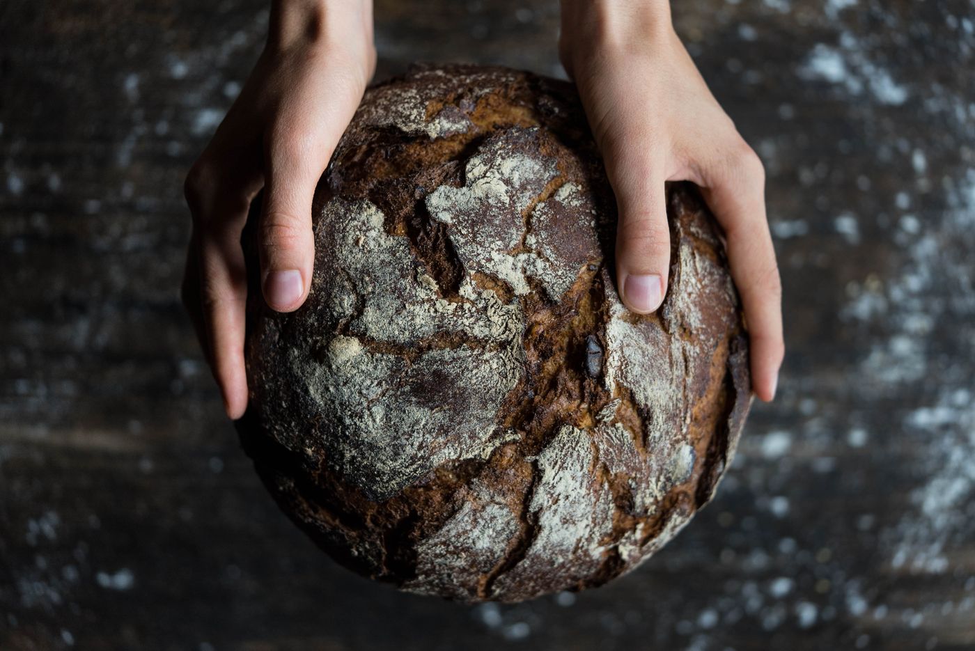 A dark, flour-dusted artisan rye loaf cradled in two hands over a rustic table