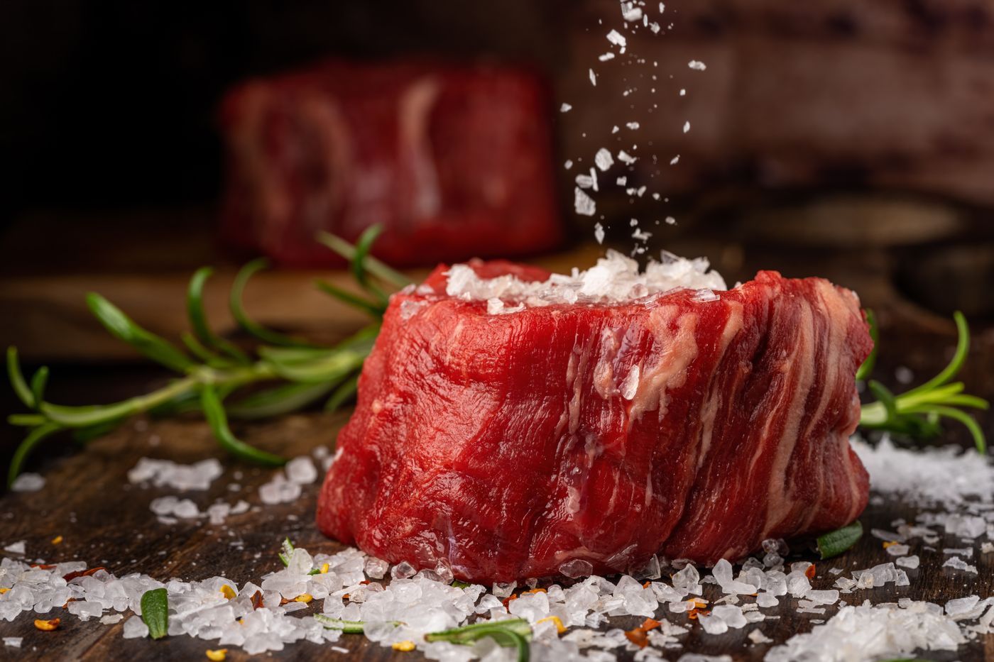 Raw steak being seasoned on a dark surface
