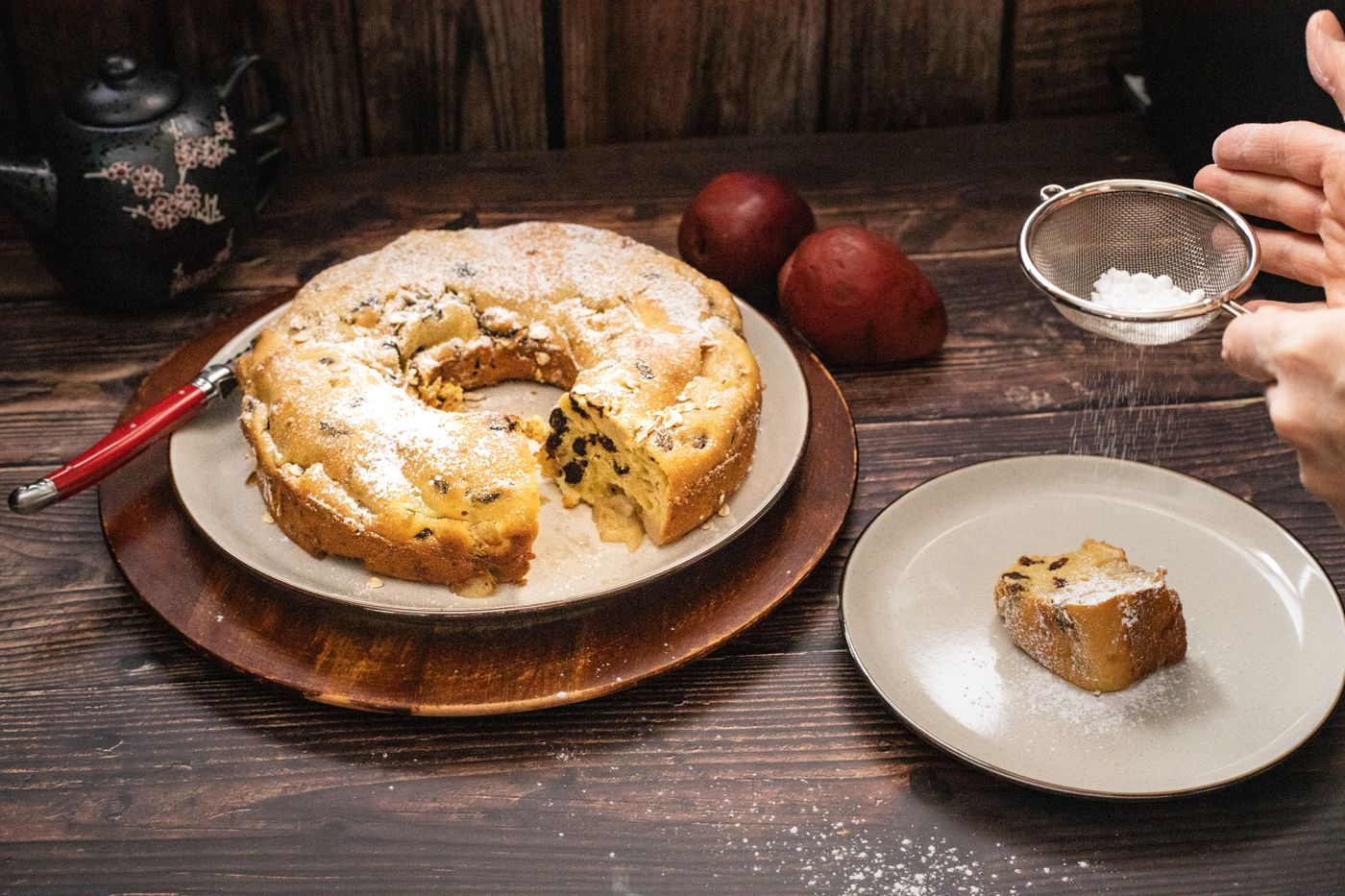 A dusted ring cake on a board, sugar being sifted over the top by hand